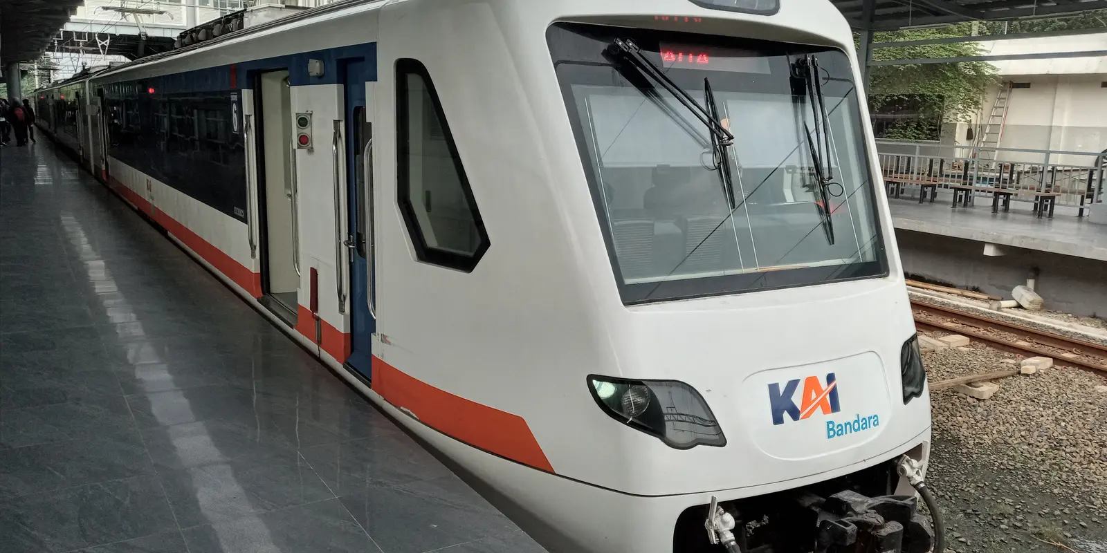 A white Soekarno-Hatta airport train with red and blue stripes and a KAI Bandara logo on its nose, standing at a covered platform at Batu Ceper station. A few passengers wait further down the platform; a second track and an opposite platform are visible to the right.